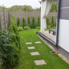 Zadbany ogród z kamiennym płotem, trawnikiem i ścieżką z płyt. Widoczny system drenażowy odprowadzający wodę z rynny.