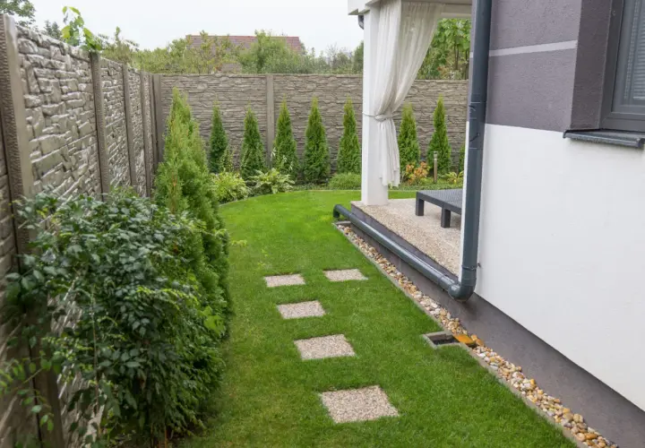 Zadbany ogród z kamiennym płotem, trawnikiem i ścieżką z płyt. Widoczny system drenażowy odprowadzający wodę z rynny.